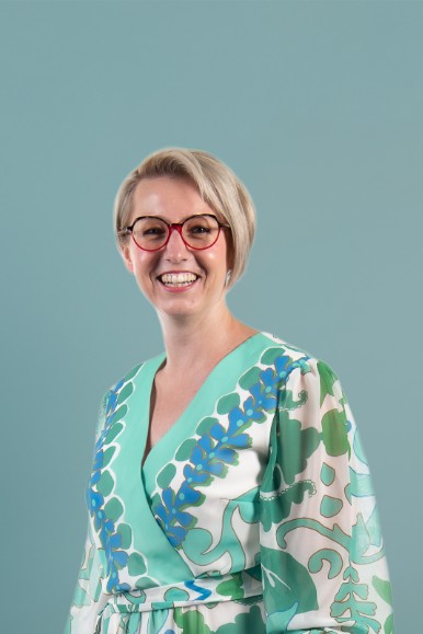 Vrouw met kort blond haar en rode bril, lachend, gekleed in een groene en witte jurk met blauw patroon. (vergroot foto)
